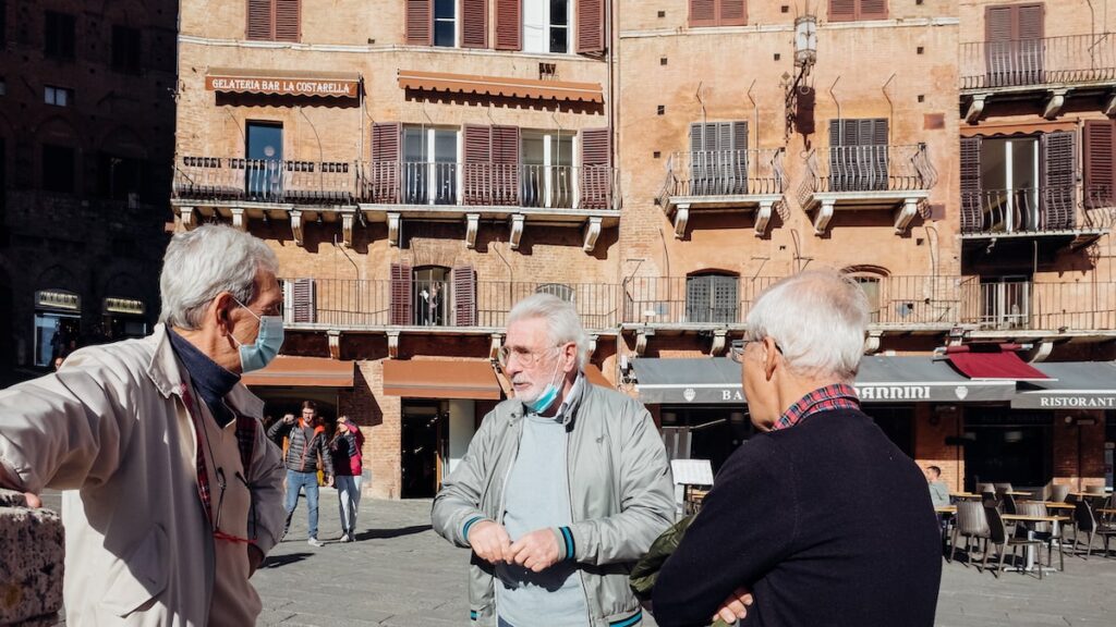 Three older men are talking in Piazza del Campo in Siena, Italy. They wear face masks due to the Covid-19 pandemic