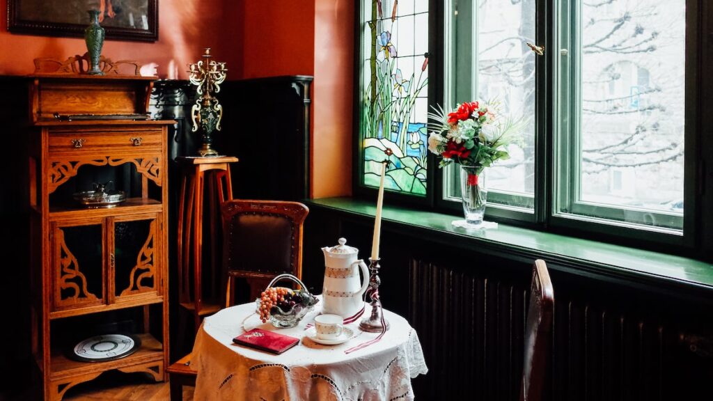 A photo from the interior of the Art Nouveau Museum in Riga, Latvia, depicting a table with a teapot, a vase with red flowers, and an ornament cupboard.