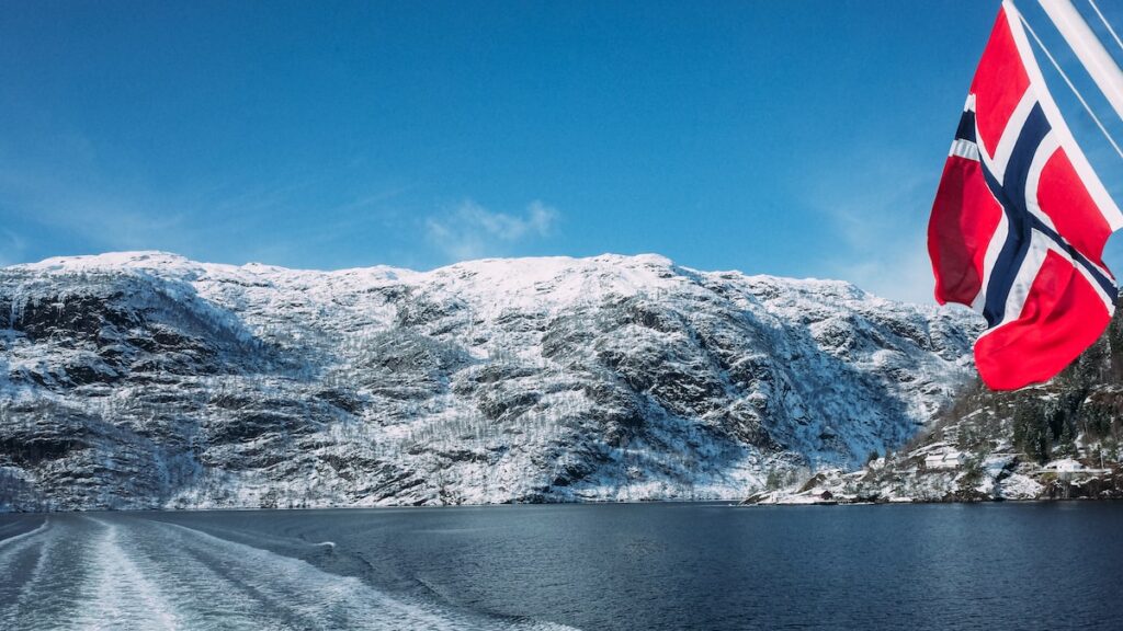 A photo depicting the snow-covered mountains of Norway. It's taken from a boat trip to Mostsraumen from Bergen, and you can see the Norwegian flag on the right.