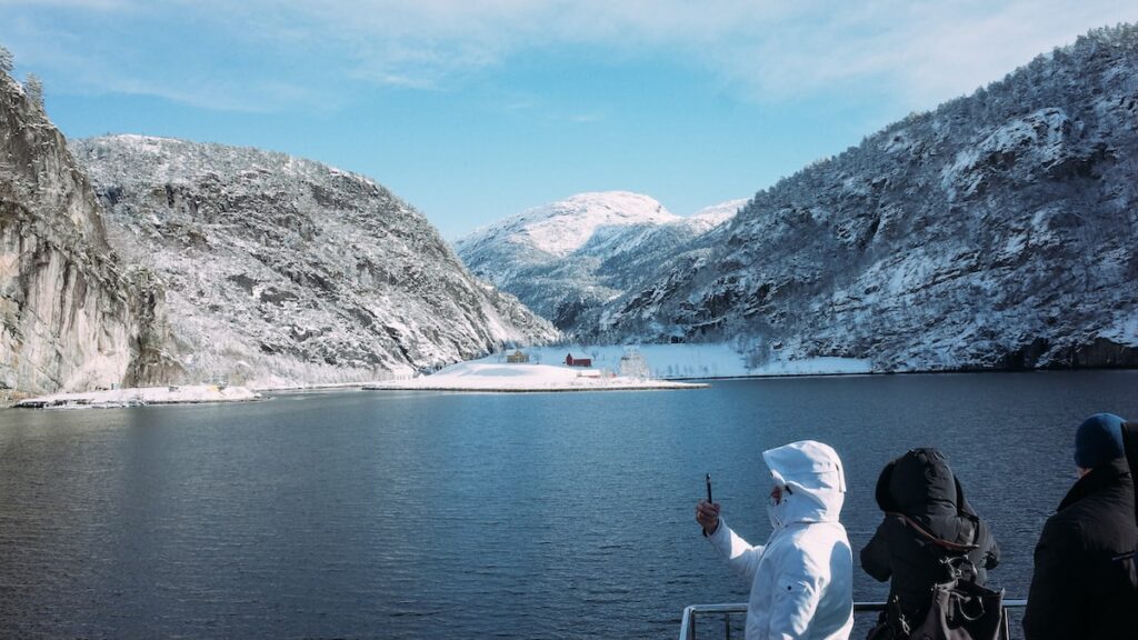 An image showing warmly dressed people and two colorful houses on the Norwegian fjords. It was taken while approaching the Mostraumen Straits on 3,5 hours fjord trip from Bergen, Norway