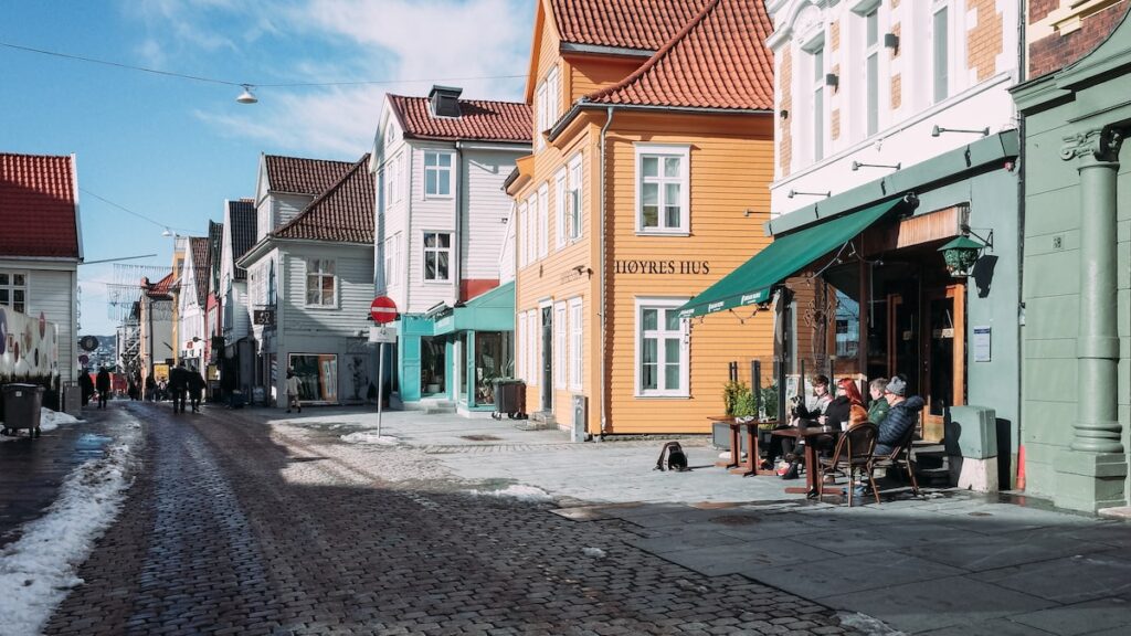An image showing a side street in Bergen with light snow, and people enjoying outdoors a cup of coffee