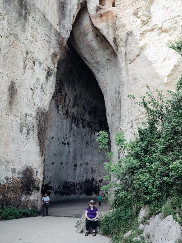 A photo of a lady sitting in front of the Ear of Dionysius in Syracuse, Italy. It is located at the Neapolis Archaeological Site and the best Syracuse tours include a visit to the site.