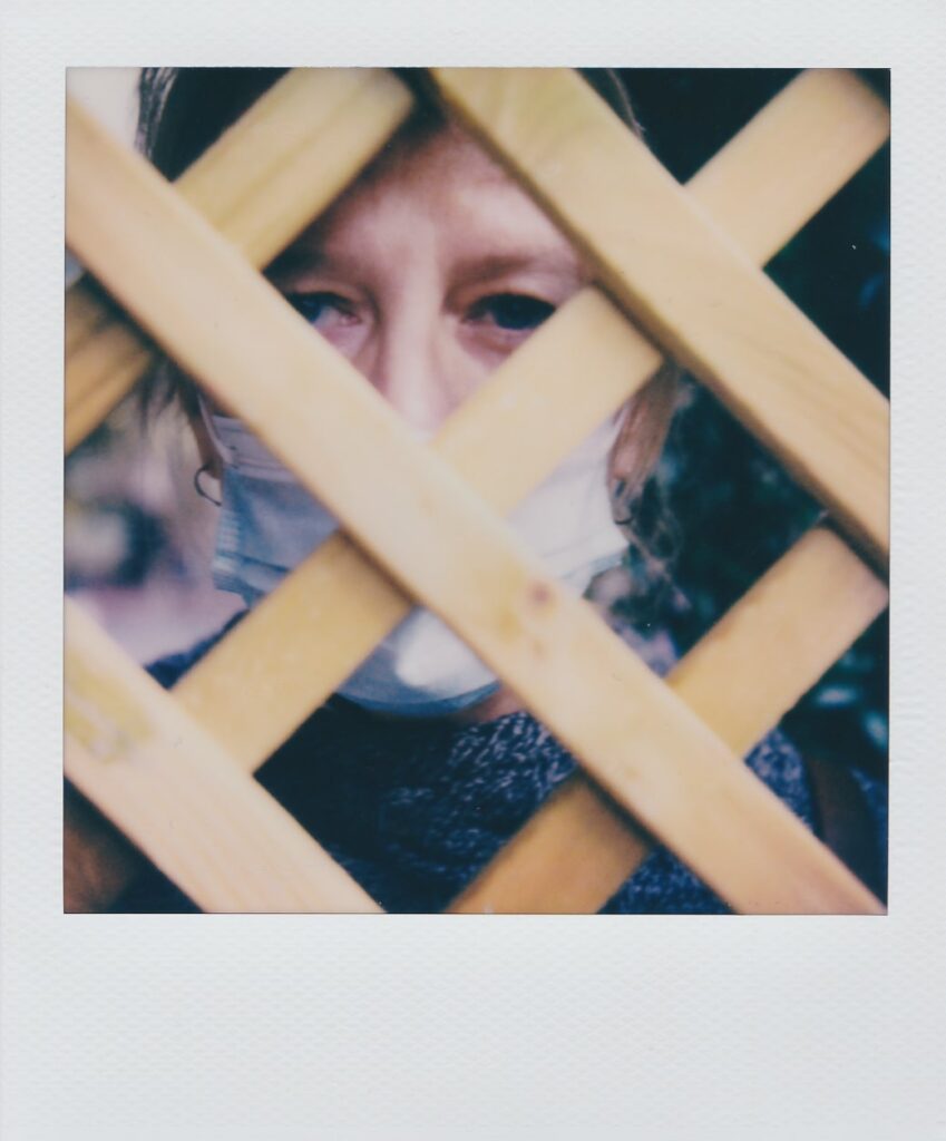 A polaroid of Barbara wearing a mask behind a fence taken with the Polaroid SX-70