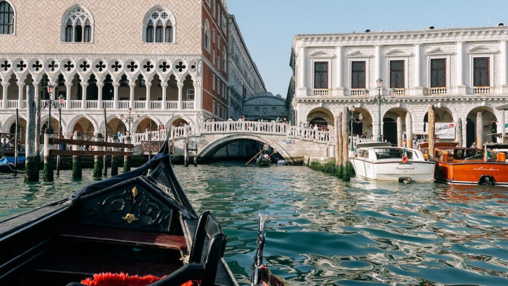 A photo showing a chair from a gondola and the city behind it. This POV photo is taken during a gondola ride in Venice and in the background you can see the Doge's Palace and the iconic Bridge of Sighs