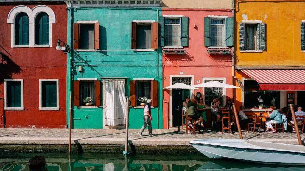 A photo of the colorful houses of Burano with people hanging out on a cafe on the right side of the photo