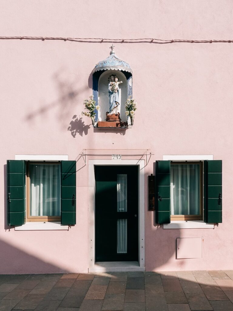A photo showing the facade of a light pink house with green windows in Burano, Italy, with a pilgrimage dedicated to Virgin Mary in the middle