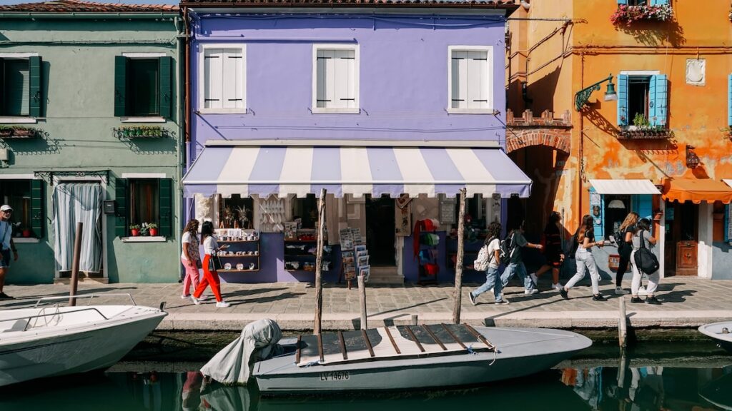A photo showing three colorful houses in Burano, Venice, while people walk in front of them and motorboats are parked in the canal