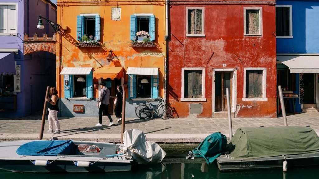 A photo showing a yellow, a red, and a blue house, with people walking in front of them in Burano, the lace island of Venice, Italy